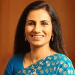A woman with long, straight, dark hair is wearing a blue saree adorned with gold patterns and delicate silver jewelry, standing against a softly lit wooden background.