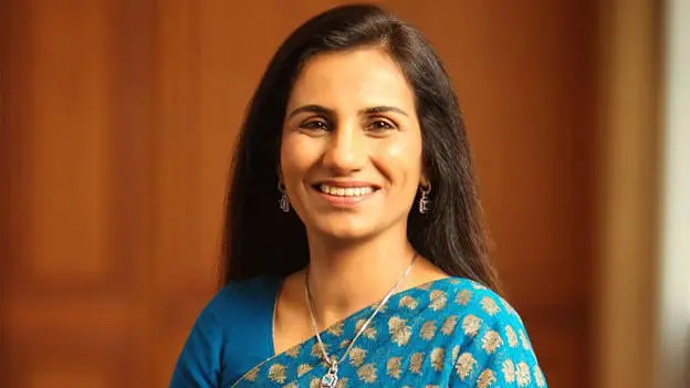 A woman with long, straight, dark hair is wearing a blue saree adorned with gold patterns and delicate silver jewelry, standing against a softly lit wooden background.